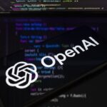 Smartphone showing OpenAI logo in front of a computer screen with code, dark background