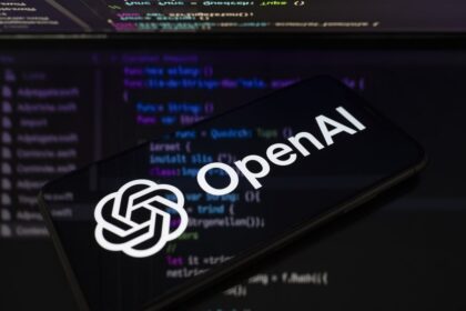 Smartphone showing OpenAI logo in front of a computer screen with code, dark background
