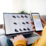 Person comparing online shopping prices on a phone with a laptop nearby