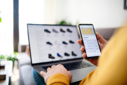 Person comparing online shopping prices on a phone with a laptop nearby